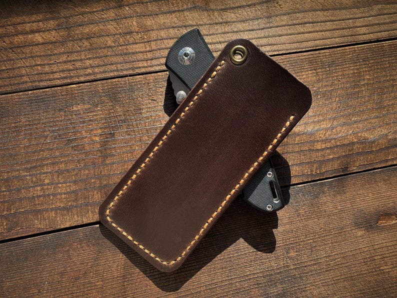 May include: A brown leather knife sheath with a contrasting stitched border and a brass grommet. The sheath is shown on a wooden surface, partially concealing a black folding knife.