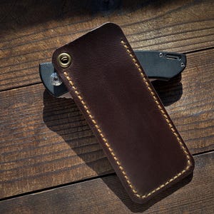 May include: A dark brown leather knife sheath with gold stitching and a brass grommet. The sheath is rectangular and covers a black and gray folding knife. The items are on a wooden surface.