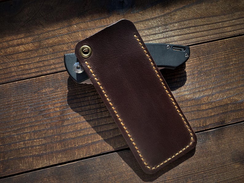 May include: A dark brown leather knife sheath with gold stitching and a brass grommet. The sheath is rectangular and covers a black and gray folding knife. The items are on a wooden surface.