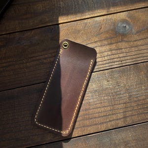 May include: A brown leather phone case with a stitched border and a brass grommet. The case is rectangular and appears to be handmade. The stitching is a light tan color, contrasting with the darker brown leather. The case is resting on a wooden surface.