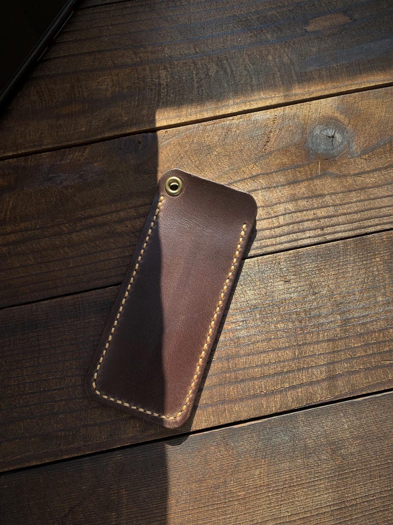 May include: A brown leather phone case with a stitched border and a brass grommet. The case is rectangular and appears to be handmade. The stitching is a light tan color, contrasting with the darker brown leather. The case is resting on a wooden surface.