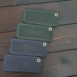May include: Four rectangular leather keychains in shades of green and blue are arranged diagonally. Each keychain has a metal eyelet and stitched edges. The word "SPOON" is embossed on the blue keychains.