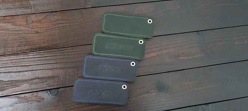 May include: Four rectangular leather keychains in shades of green and blue are arranged diagonally. Each keychain has a metal eyelet and stitched edges. The word "SPOON" is embossed on the blue keychains.