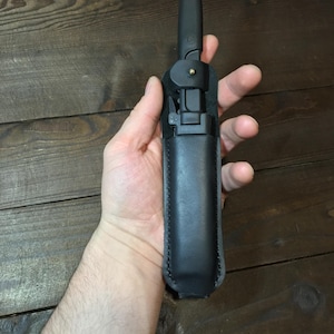 May include: A black knife with a black leather sheath. The knife is partially extended and held in place by a metal clasp. The sheath is made of stitched leather. The knife and sheath are on a dark wood surface.