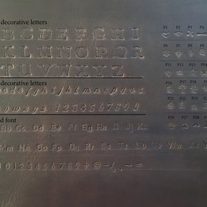 May include: A gray metal surface with embossed lettering and symbols. The image displays large and small decorative letters, a printed font, numbers, and various symbols, including hearts, stars, and crosses.