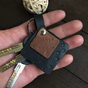 Puede incluir: Un llavero de cuero negro con una etiqueta de cuero marrón que dice "Mushyn Roman" con huellas de patas. El llavero tiene un anillo de metal plateado y está unido a un juego de llaves.
