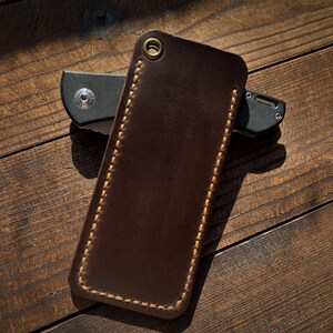 May include: A brown leather knife sheath with a contrasting stitched border and a brass grommet. A black folding knife is partially visible behind the sheath. The items are on a wooden surface.