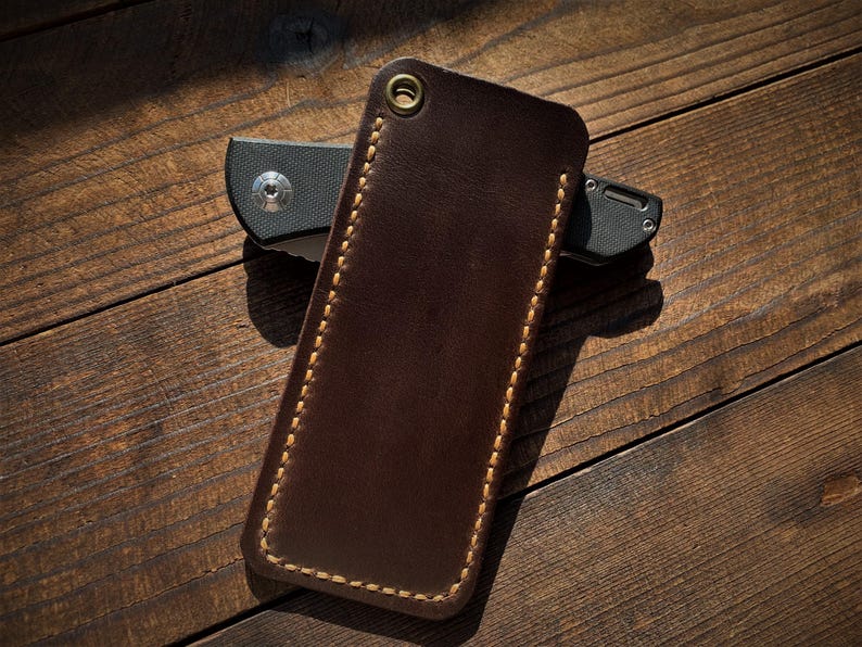 May include: A brown leather knife sheath with a contrasting stitched border and a brass grommet. A black folding knife is partially visible behind the sheath. The items are on a wooden surface.