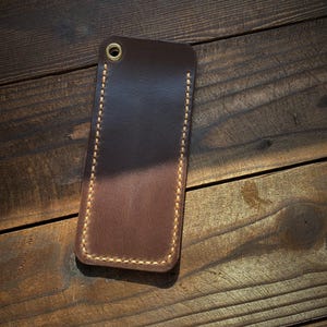 May include: A rectangular, brown leather phone case with a stitched border and a brass grommet at the top. The case is set against a dark wooden background, showcasing its handcrafted details.