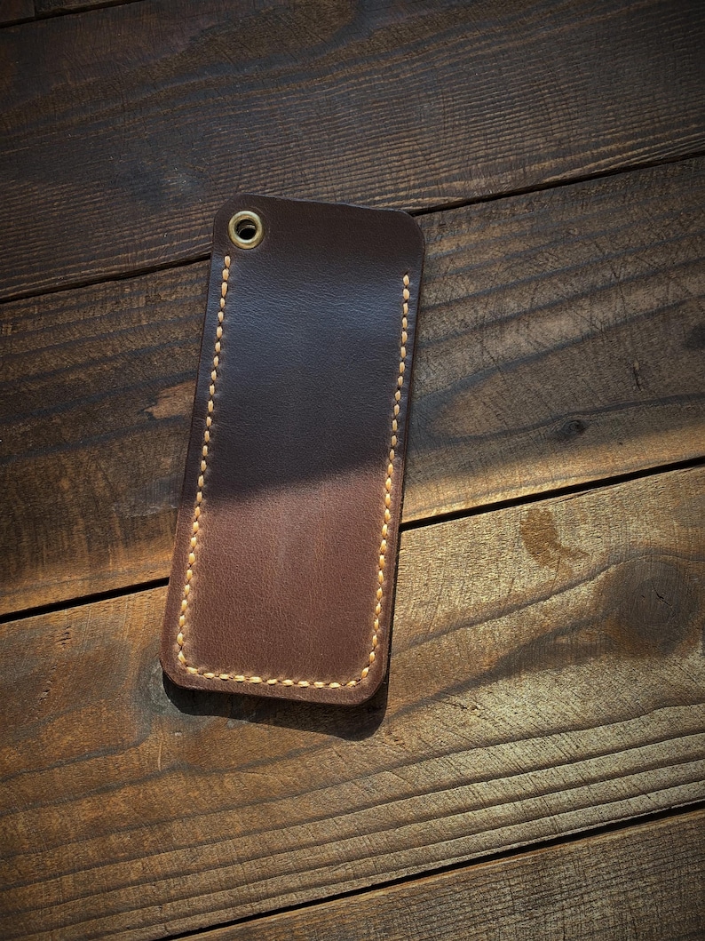 May include: A rectangular, brown leather phone case with a stitched border and a brass grommet at the top. The case is set against a dark wooden background, showcasing its handcrafted details.