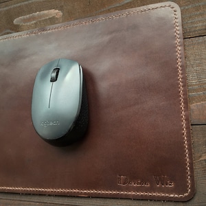 Puede incluir: Una alfombrilla de ratón de cuero marrón con un borde cosido y el nombre "Daria Wiz" grabado en la esquina inferior derecha. Un ratón inalámbrico plateado está en la alfombrilla.