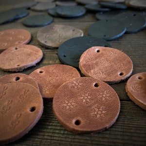Può includere: Una collezione di etichette in pelle marrone con vari disegni, tra cui motivi floreali e note musicali. Le etichette hanno due fori ciascuna e sono disposte su una superficie di legno.