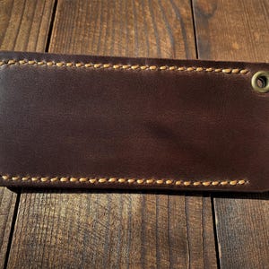 May include: A rectangular, dark brown leather item with a stitched border and a brass grommet. The leather has a smooth surface and a contrasting light brown stitching. The item is likely a wallet or card holder.