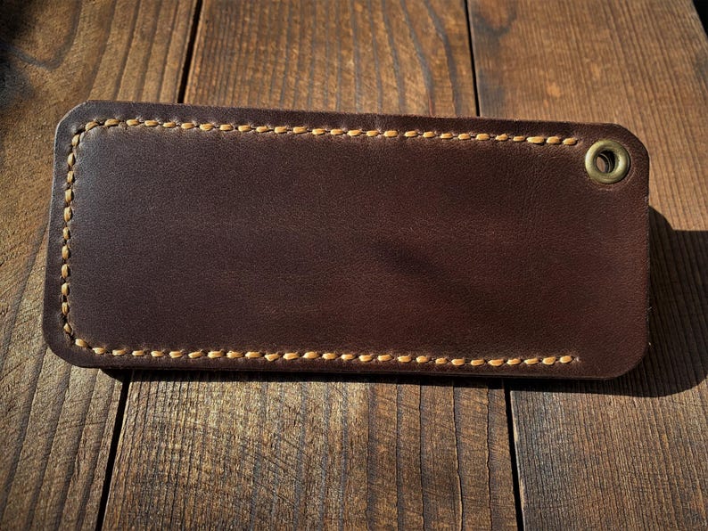 May include: A rectangular, dark brown leather item with a stitched border and a brass grommet. The leather has a smooth surface and a contrasting light brown stitching. The item is likely a wallet or card holder.