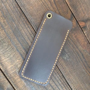 May include: A dark brown leather key fob holder with a stitched border and a brass grommet. The rectangular case is shown on a wooden surface.