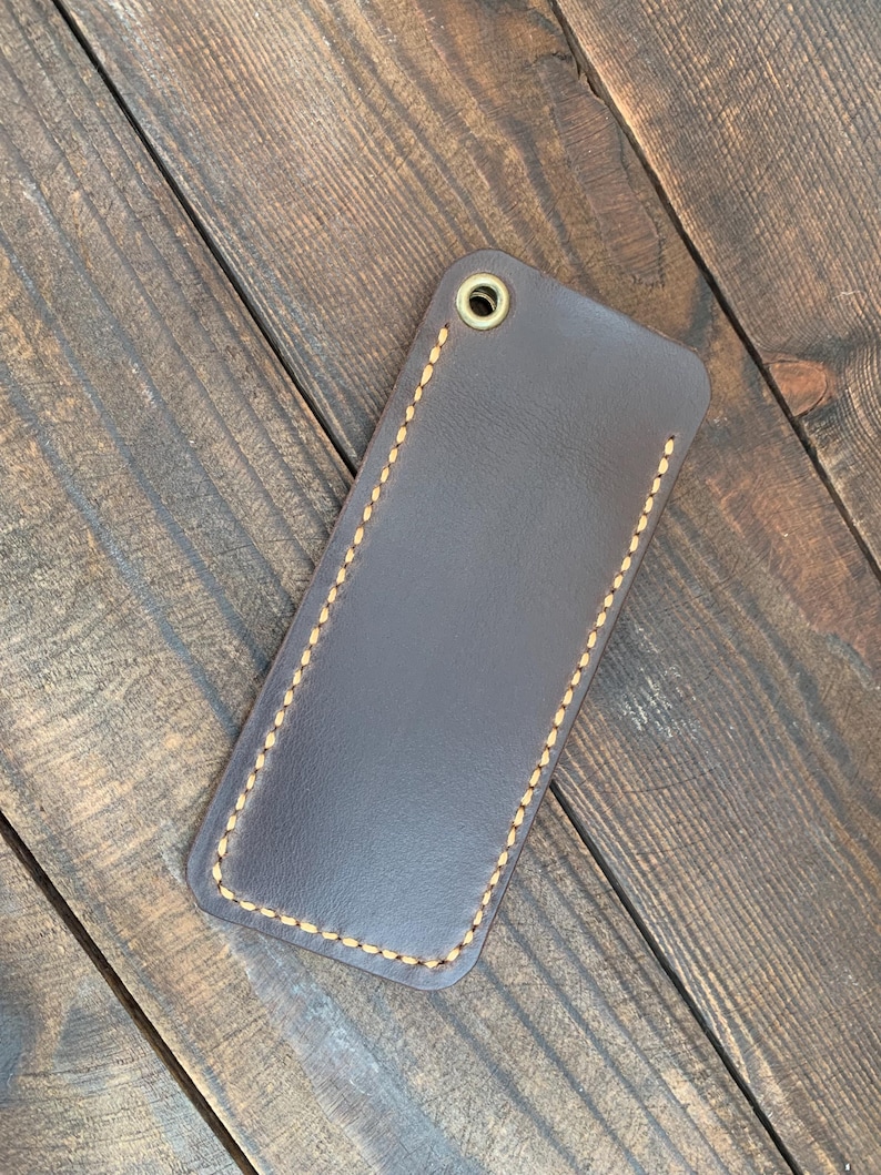 May include: A dark brown leather key fob holder with a stitched border and a brass grommet. The rectangular case is shown on a wooden surface.