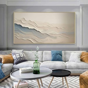 May include: A large, rectangular abstract artwork in a gold frame. The piece depicts layered, flowing forms in beige, white, and blue, reminiscent of a landscape. It hangs above a grey sofa with decorative cushions.