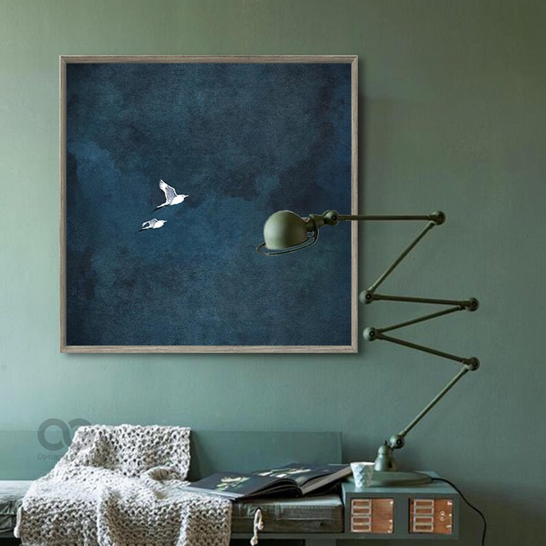 May include: A framed artwork depicting two white seagulls soaring against a dark blue, textured backdrop. The picture hangs above a green sofa, with a blanket and a green desk lamp visible.