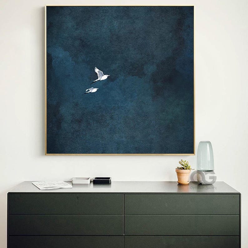 May include: A framed print depicting two white birds soaring against a dark blue, textured backdrop. The artwork, in a gold frame, hangs above a dark grey chest of drawers.