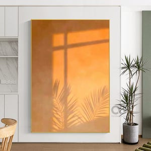 May include: An abstract framed artwork with a warm orange and yellow gradient background. The artwork features shadows of palm leaves and a window frame, creating a natural and serene ambiance. The artwork is displayed in a bright, modern interior.