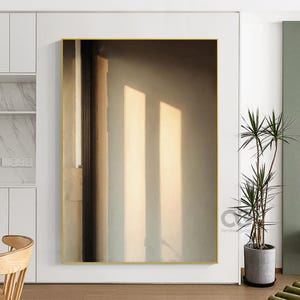 Pode incluir: Arte de parede abstrata com moldura dourada, apresentando uma composição vertical em tons quentes e neutros. A obra captura a interação de luz e sombra, oferecendo uma estética minimalista. Uma planta em vaso é visível no canto.
