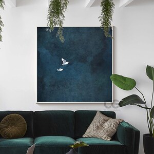 May include: A framed art print showing two white seagulls soaring against a deep blue, textured backdrop. The artwork is displayed above a dark green velvet sofa. The print is square.