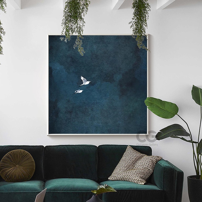May include: A framed art print showing two white seagulls soaring against a deep blue, textured backdrop. The artwork is displayed above a dark green velvet sofa. The print is square.