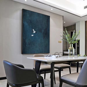 May include: A framed canvas artwork featuring two white birds soaring against a dark teal backdrop. The painting hangs on a light grey wall above a dining table and chairs. The room has a modern, minimalist design with a vase of greenery.