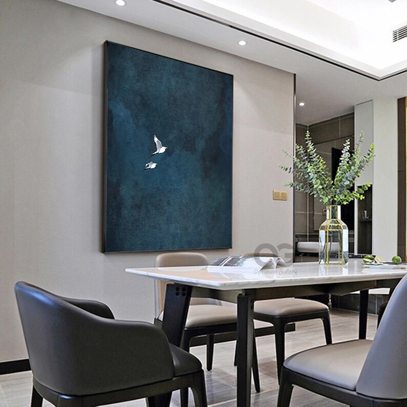 May include: A framed canvas artwork featuring two white birds soaring against a dark teal backdrop. The painting hangs on a light grey wall above a dining table and chairs. The room has a modern, minimalist design with a vase of greenery.