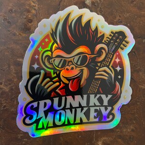 May include: A holographic sticker featuring a cartoon monkey with sunglasses and a mohawk playing a guitar. The monkey is wearing a red jacket and has a tongue sticking out. The text "SPUNKY MONKEY" is written below the monkey.