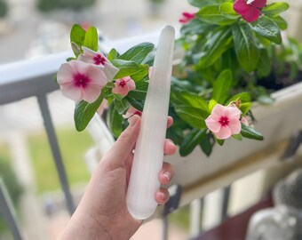 Puede incluir: Una varita de selenita blanca, también conocida como varita de limpieza, se sostiene en una mano. La varita es lisa y pulida. El fondo presenta flores rosas.