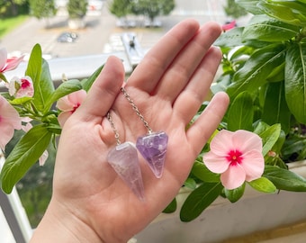Puede incluir: Dos péndulos de cristal de amatista en cadenas plateadas, sostenidos en una mano sobre un fondo de plantas verdes y flores rosas.