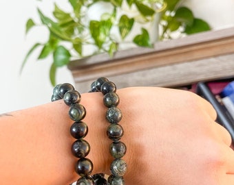 May include: Two bracelets made of dark green and black gemstone beads. The bracelets are stacked on a person's wrist.