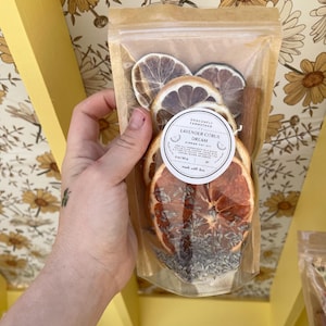 Può includere: Un sacchetto trasparente pieno di fette di agrumi essiccati, un bastoncino di cannella e lavanda. L'etichetta circolare recita "Lavender Citrus Dream Simmer Pot Kit". Il sacchetto è tenuto davanti a una carta da parati floreale.