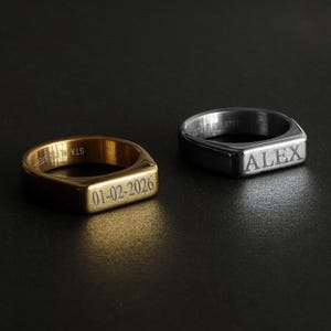 May include: Two stainless steel signet rings. One is gold-toned with the date "01-02-2026" engraved. The other is silver-toned with the name "ALEX" engraved. Both rings have a rectangular face and a polished finish.