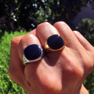 May include: Two signet rings featuring smooth, rounded square black stone inlays. One ring is crafted from polished silver-toned metal, while the other is made from polished gold-toned metal. Both rings have a solid, wide band design.
