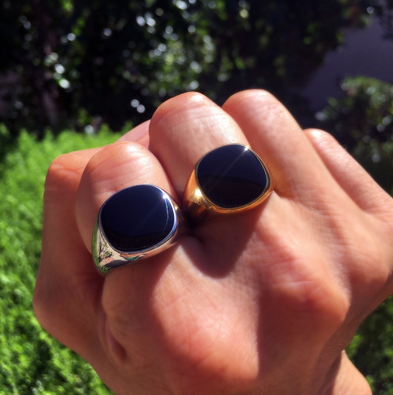 May include: Two signet rings featuring smooth, rounded square black stone inlays. One ring is crafted from polished silver-toned metal, while the other is made from polished gold-toned metal. Both rings have a solid, wide band design.