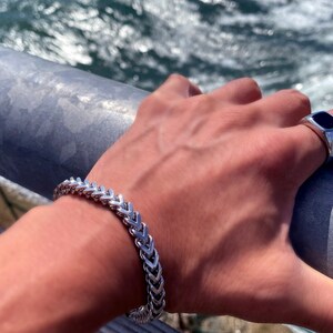 Puede incluir: Un brazalete de cadena de plata con un dise&ntilde;o de cola de pez. El brazalete est&aacute; en la mu&ntilde;eca de una persona. La persona tambi&eacute;n lleva un anillo de plata con una piedra negra.