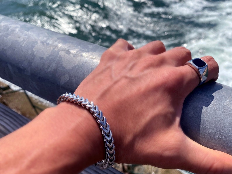 Puede incluir: Un brazalete de cadena de plata con un dise&ntilde;o de cola de pez. El brazalete est&aacute; en la mu&ntilde;eca de una persona. La persona tambi&eacute;n lleva un anillo de plata con una piedra negra.