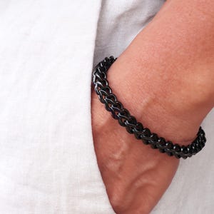May include: A polished black metal chain link bracelet featuring a series of interconnected, rounded oval links. The bracelet has a smooth, reflective surface and a consistent dark finish throughout the entire length of the linked chain design.