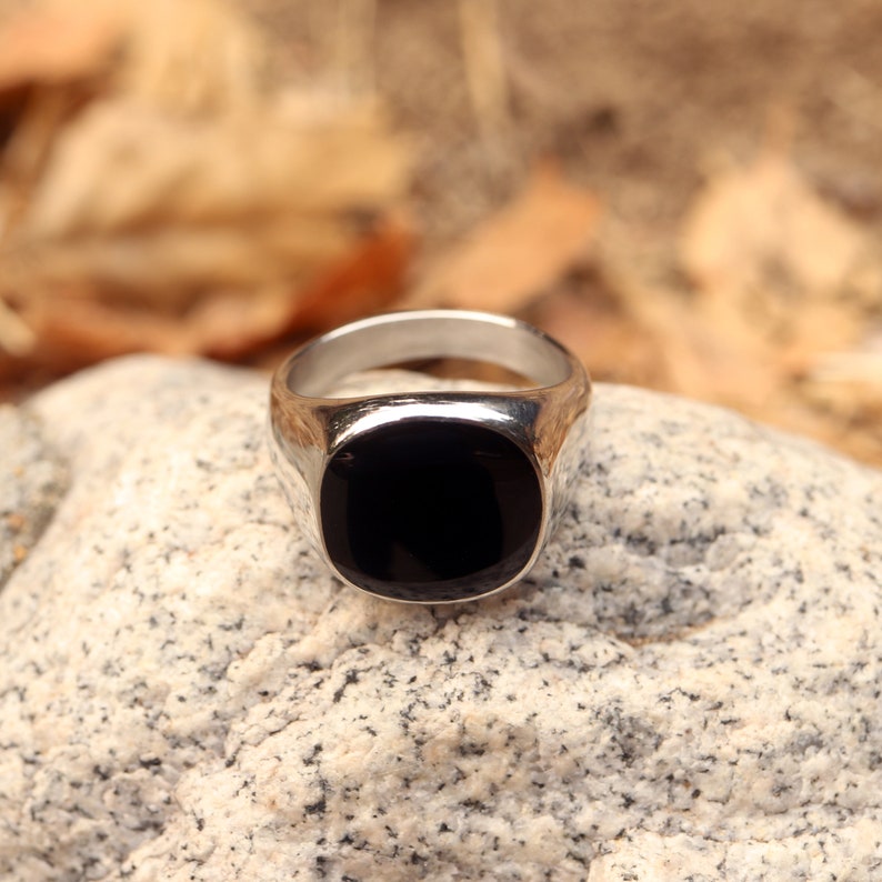May include: A silver signet ring featuring a smooth, polished black stone inlay. The ring has a wide, rounded band that tapers toward the back, with a flat, square-shaped face that holds the dark, reflective stone securely in the center.