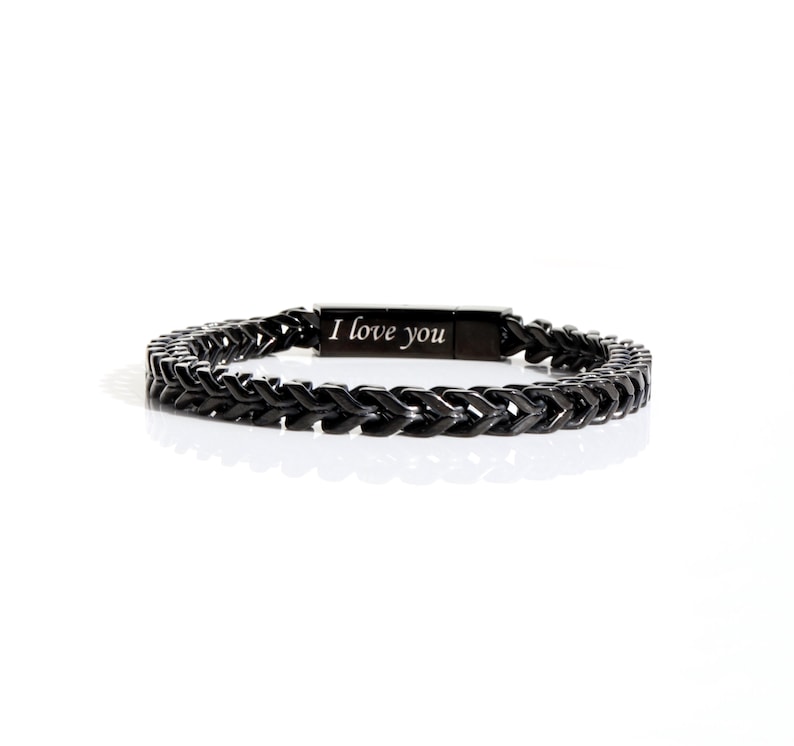 May include: A black metal bracelet featuring a woven chain link design and a rectangular clasp engraved with the text I love you. The bracelet has a dark, polished finish and a structured, repeating pattern throughout the entire band.