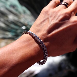 May include: A silver-toned metal chain bracelet featuring a woven wheat link pattern. The bracelet has a polished, reflective surface and a rounded, substantial profile that wraps around the wrist, showcasing a consistent interlocking design.