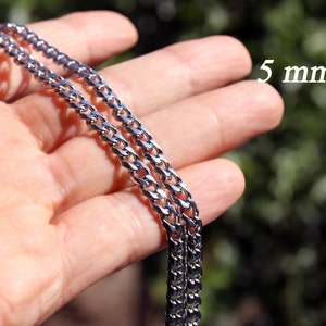 K&ouml;nnte beinhalten: Eine silberne Curb-Kette mit einer Breite von 5 mm. Die Kette wird auf einer Hand gezeigt, wobei die Breite der Kette durch den Text "5 mm" angezeigt wird.