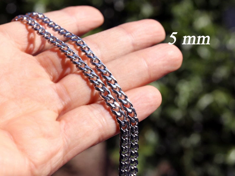 K&ouml;nnte beinhalten: Eine silberne Curb-Kette mit einer Breite von 5 mm. Die Kette wird auf einer Hand gezeigt, wobei die Breite der Kette durch den Text "5 mm" angezeigt wird.