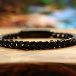 May include: A black bracelet featuring a continuous wheat chain design with interlocking metal links. The bracelet has a smooth, dark finish and a rectangular clasp component visible at the center, creating a uniform and structured appearance.