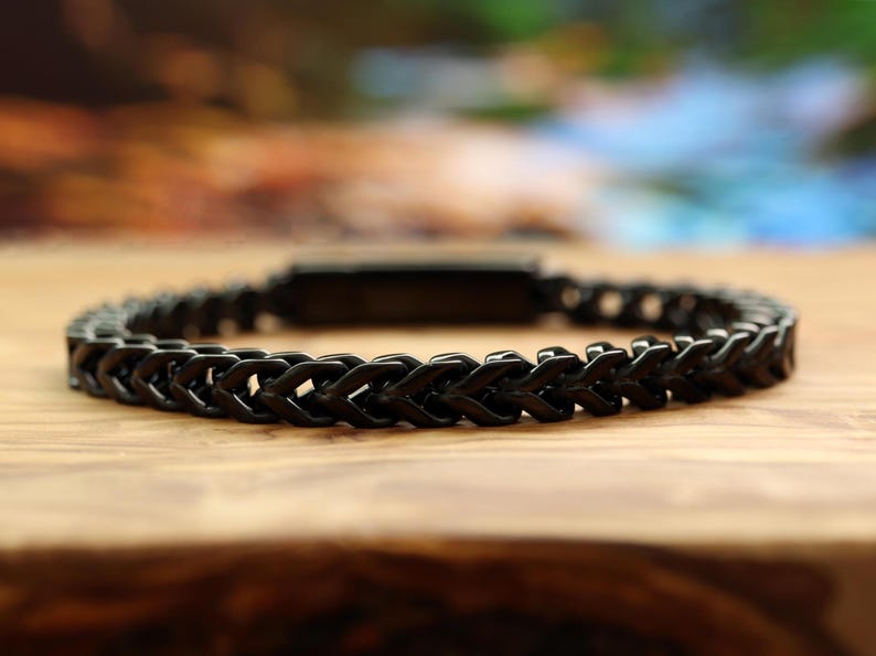 May include: A black bracelet featuring a continuous wheat chain design with interlocking metal links. The bracelet has a smooth, dark finish and a rectangular clasp component visible at the center, creating a uniform and structured appearance.