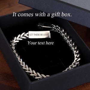 May include: A silver-toned wheat chain bracelet with a rectangular metal clasp featuring the engraved text "LET THERE BE LIGHT" and "Your text here," presented inside a black gift box with the printed text "It comes with a gift box."