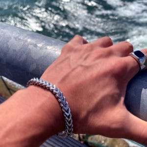 May include: A silver-toned metal bracelet featuring a repeating chevron or wheat-link pattern, paired with a silver-toned signet ring that has a smooth, rectangular black inlay on the top surface, both worn on the wrist and finger.