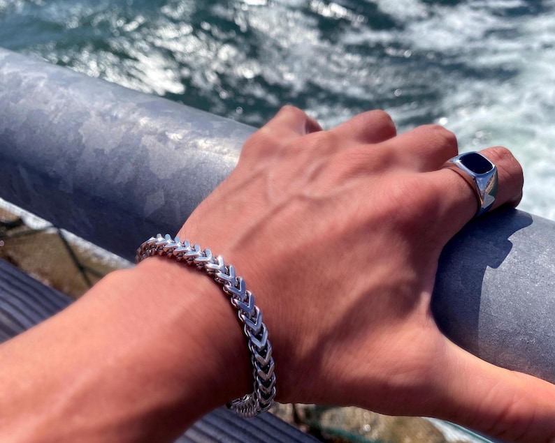 May include: A silver-toned metal bracelet featuring a repeating chevron or wheat-link pattern, paired with a silver-toned signet ring that has a smooth, rectangular black inlay on the top surface, both worn on the wrist and finger.